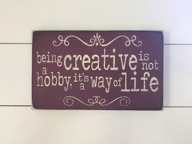 May include: A barn red wooden sign with cream text that reads "being creative is not a hobby, it's a way of life". The sign has decorative swirls at the top and bottom.