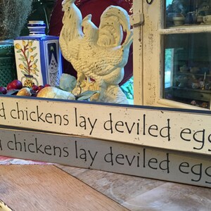 May include: Two wooden signs with the text "Wicked chickens lay deviled eggs" in white lettering. The signs are stacked on top of each other, with the top sign being a light beige color and the bottom sign being a light gray color.