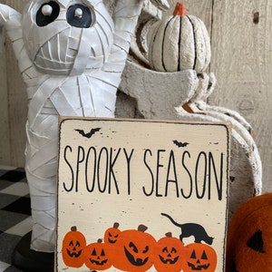 May include: A square, off-white wooden sign with the words "Spooky Season" in black lettering. Below the text are orange jack-o'-lanterns and a black cat silhouette. The sign is propped up on a black metal stand, with a white mummy statue in the background.