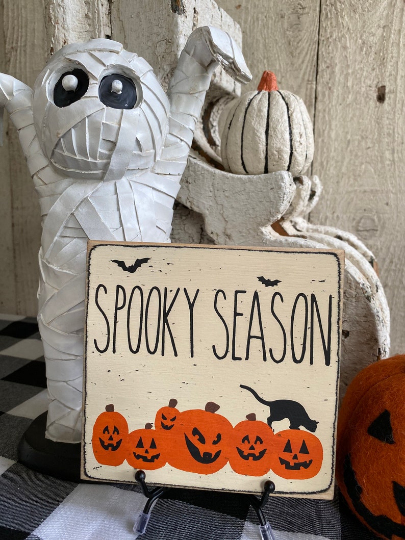 May include: A square, off-white wooden sign with the words "Spooky Season" in black lettering. Below the text are orange jack-o'-lanterns and a black cat silhouette. The sign is propped up on a black metal stand, with a white mummy statue in the background.