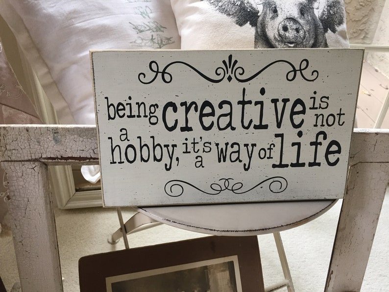 May include: White wooden sign with black text that reads "being creative is not a hobby, it's a way of life". The sign has decorative swirls at the top and bottom.