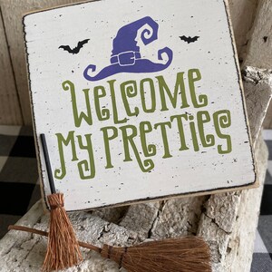 May include: A square, white wooden sign with the text "Welcome My Pretties" in green lettering, a purple witch hat, and two black bats. Two small brown brooms are in front of the sign. The sign has a distressed, vintage look.
