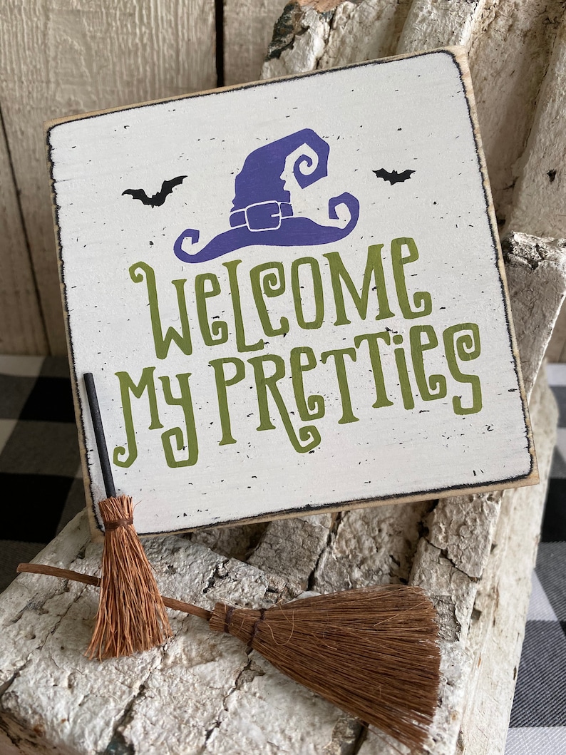 May include: A square, white wooden sign with the text "Welcome My Pretties" in green lettering, a purple witch hat, and two black bats. Two small brown brooms are in front of the sign. The sign has a distressed, vintage look.