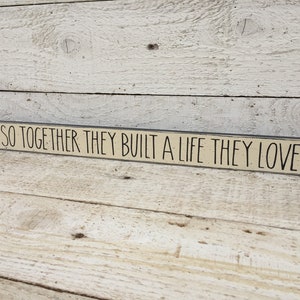 May include: A wooden sign with black text that reads "And so together they built a life they loved".