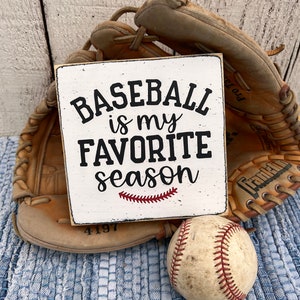 Op de afbeelding: Een wit houten bord met zwarte tekst die "Baseball is my favorite season" luidt, met een rood gestikt honkbalnaadontwerp. Op de achtergrond zijn een bruine honkbalhandschoen en een witte honkbal te zien.
