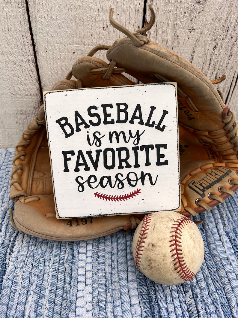 Op de afbeelding: Een wit houten bord met zwarte tekst die "Baseball is my favorite season" luidt, met een rood gestikt honkbalnaadontwerp. Op de achtergrond zijn een bruine honkbalhandschoen en een witte honkbal te zien.