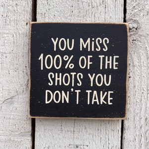 May include: Black wooden sign with white text that reads "You miss 100% of the shots you don't take."
