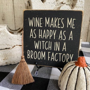 May include: A black wooden sign with white lettering that reads "WINE MAKES ME AS HAPPY AS A WITCH IN A BROOM FACTORY." A small broom and a white pumpkin with black stripes are also in the image. The background is a black and white checkered pattern.