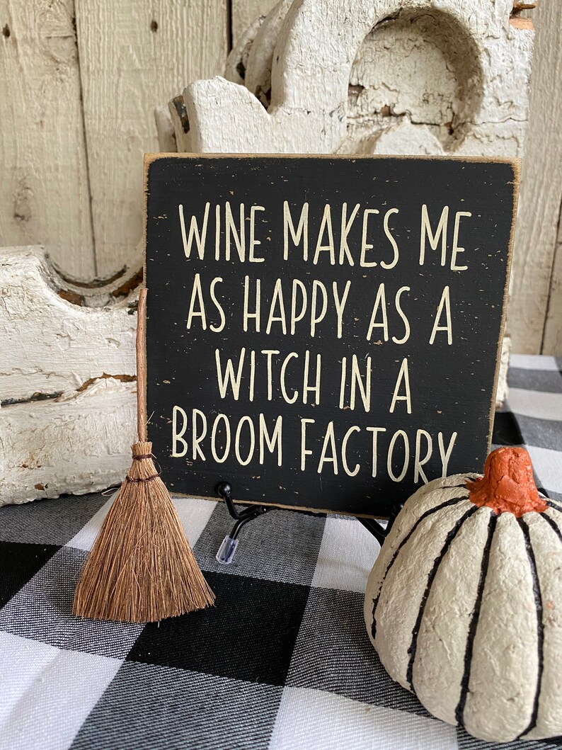 May include: A black wooden sign with white lettering that reads "WINE MAKES ME AS HAPPY AS A WITCH IN A BROOM FACTORY." A small broom and a white pumpkin with black stripes are also in the image. The background is a black and white checkered pattern.