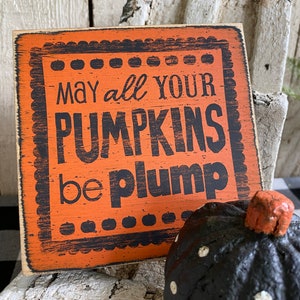 May include: Orange wooden sign with the text "May all your pumpkins be plump" in black lettering. A black pumpkin with white polka dots and an orange stem is next to the sign. The sign has a decorative border.