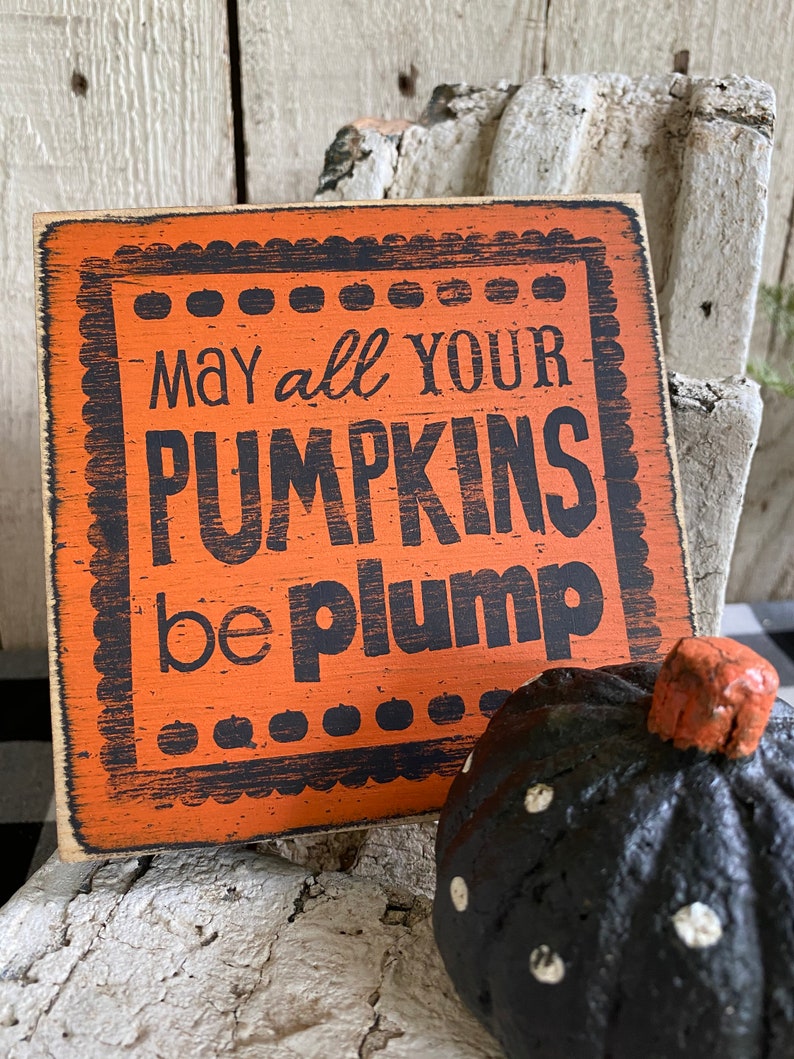 May include: Orange wooden sign with the text "May all your pumpkins be plump" in black lettering. A black pumpkin with white polka dots and an orange stem is next to the sign. The sign has a decorative border.