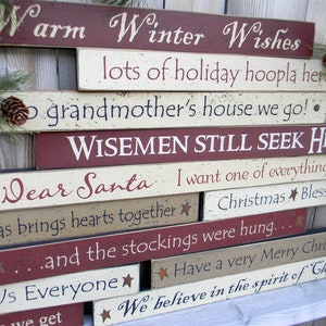 May include: A collection of rustic wooden signs with Christmas-themed phrases in white lettering on a red, beige, and cream background. Phrases include "Warm Winter Wishes" and "Dear Santa". The signs are decorated with stars.