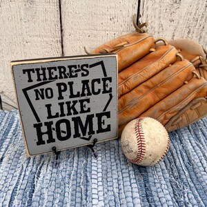 Op de afbeelding: Een grijs houten bord met zwarte tekst die "There's No Place Like Home" luidt. Het bord staat op een blauw-wit gestreept tapijt met een honkbal en een bruine leren honkbalhandschoen.