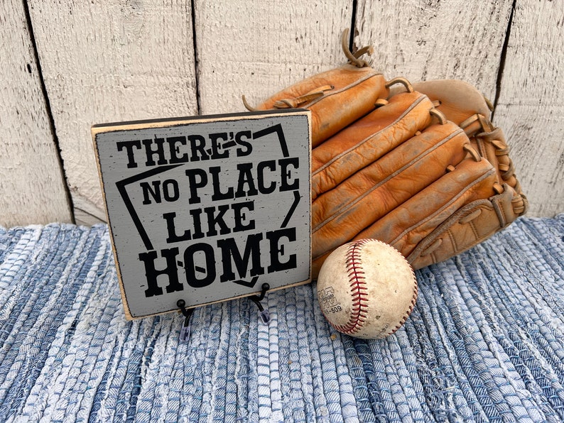 Op de afbeelding: Een grijs houten bord met zwarte tekst die "There's No Place Like Home" luidt. Het bord staat op een blauw-wit gestreept tapijt met een honkbal en een bruine leren honkbalhandschoen.