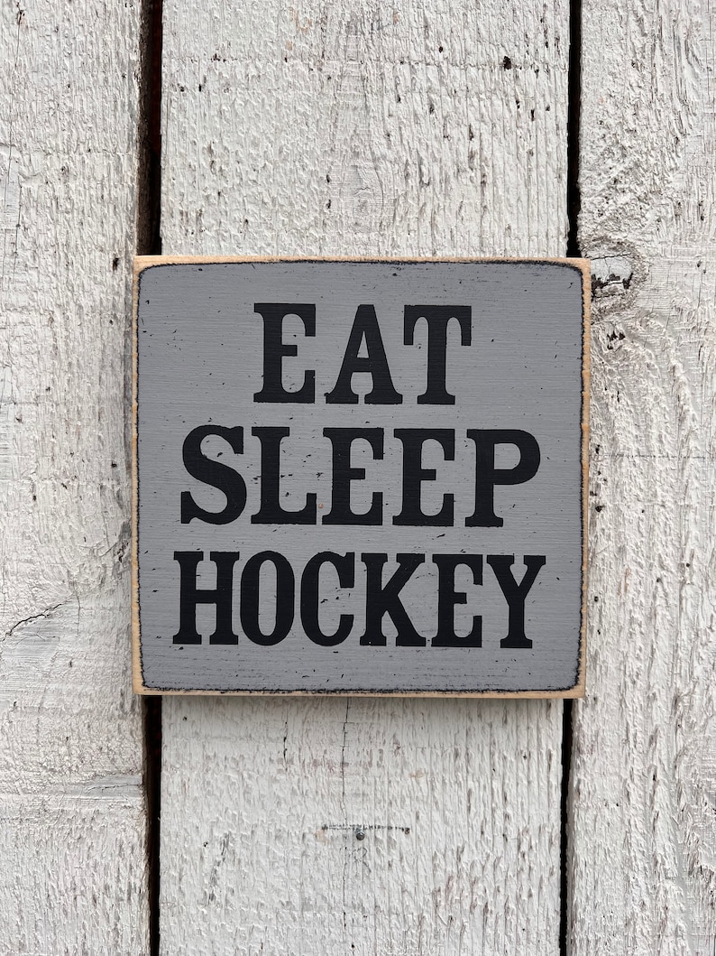 May include: A gray wooden sign with black text that reads "Eat Sleep Hockey".
