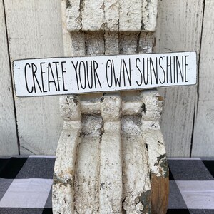 May include: White wooden sign with black text that reads "CREATE YOUR OWN SUNSHINE". The sign is sitting on a white, weathered, wooden corbel.