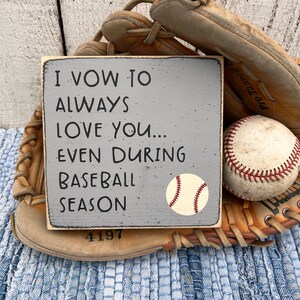 Op de afbeelding: Een grijs houten bord met de tekst "I vow to always love you... even during baseball season" en een baseball afbeelding. Het bord ligt op een blauw-wit geweven tapijt met een honkbalhandschoen en een honkbal.