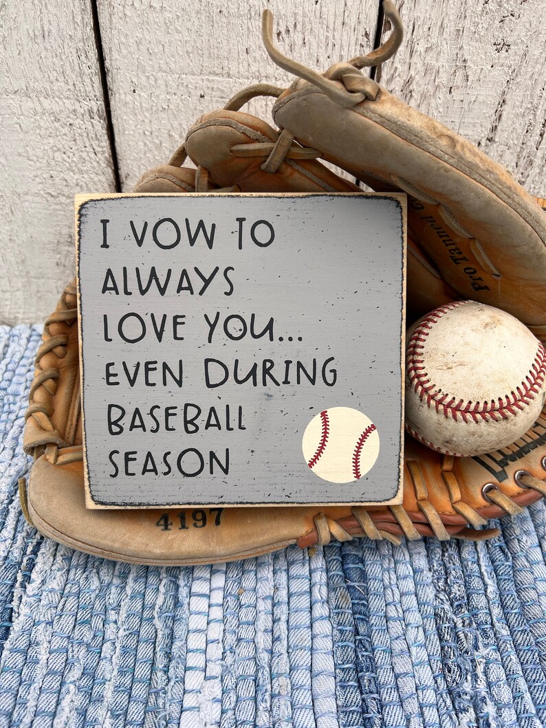 Op de afbeelding: Een grijs houten bord met de tekst "I vow to always love you... even during baseball season" en een baseball afbeelding. Het bord ligt op een blauw-wit geweven tapijt met een honkbalhandschoen en een honkbal.