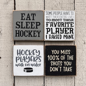 May include: Four wooden signs with hockey-themed sayings. The signs are painted in black, white, and gray. The text on the signs reads: "Eat Sleep Hockey", "Some people have to wait their entire lives to meet their favorite player. I raised mine", "Hockey players walk on water", and "You miss 100% of the shots you don't take".