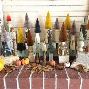 May include: A collection of decorative bottle brush trees in various colors, including orange, yellow, and green, displayed on pedestals. Miniature pumpkins and fall leaves are scattered around the base, creating a seasonal autumn theme.