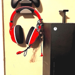 Pode incluir: Um console Xbox preto com um logotipo branco, um fone de ouvido para jogos vermelho e um controle Xbox preto montado na parede.