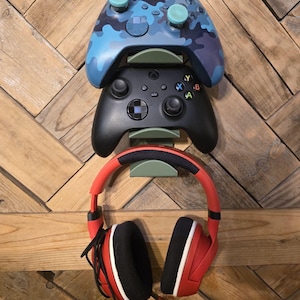 May include: A wall-mounted gaming headset holder with two video game controllers and a pair of red gaming headphones. The controllers are black and blue with camouflage patterns and have blue and green buttons. The headphones have black earcups with red accents.