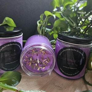 May include: Three purple soy candles with black lids and labels that say "hand-crafted with herbs & essential oils" and "SPLIT TONGUE AS NIA" with a natural soy candle logo. The candles are on a wooden surface with green leafy plants.