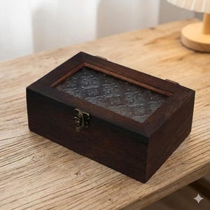 May include: A rectangular wooden box with a dark brown finish. The box features a clear glass top with a decorative floral pattern. A metal clasp secures the lid, and the box is set on a wooden surface.