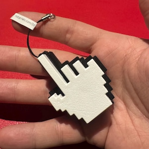 May include: A white and black pixelated cursor charm with a black cord and silver clasp. The charm is held in a hand. The tag reads rodyprieto.com/3d.