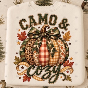 May include: A cream-colored sweatshirt featuring a central graphic of a pumpkin with segments in camouflage, leopard print, red gingham, and floral patterns. A camouflage bow sits atop the pumpkin. The text reads CAMO & cozy with autumn leaves.