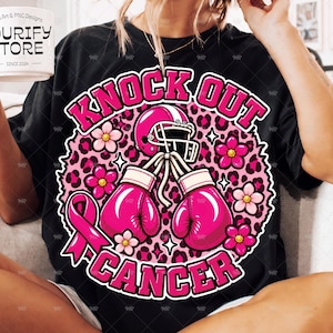 Może przedstawiać: Czarny t-shirt z grafiką zawierającą napis "KNOCK OUT CANCER" różowymi literami. Wzór przedstawia różowy kask do futbolu amerykańskiego, dwie różowe rękawice bokserskie, różową wstążkę oraz różowe kwiaty na tle w różowy wzór panterki.