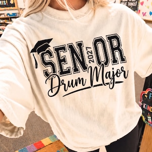 May include: An off-white short-sleeve t-shirt featuring black text that reads SENIOR 2027 Drum Major. The design includes a black graduation cap icon positioned above the word SENIOR, with the year 2027 printed vertically between the words.
