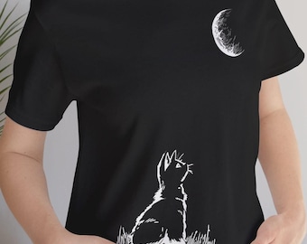 May include: Black t-shirt featuring a white illustration of a cat gazing at a crescent moon. The cat is sitting in grass, and the moon is in the upper right corner. The shirt is made of a soft material.