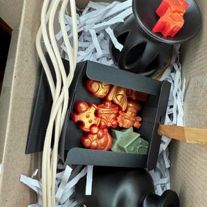 Puede incluir: Una caja de regalo llena de fundidos de cera perfumados negros y dorados, un quemador de cera de cerámica negra y palitos difusores de madera. La caja está llena de papel triturado.