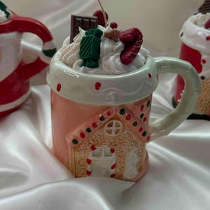 Puede incluir: Una vela en forma de taza de cerámica rosa con un diseño de casa de jengibre. La vela está cubierta con glaseado blanco, chocolate y decoraciones de caramelos. La taza tiene un asa y un borde blancos con detalles rojos.