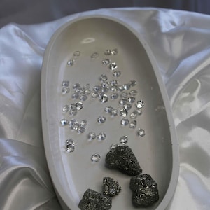 Op de afbeelding: Een witte ovale keramische schaal met een strooiing van kleine, heldere glazen stenen en vier ruwe, donkergrijze stenen.