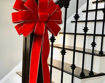 May include: A large, vibrant red velvet bow with a metallic gold-trimmed edge, tied securely to the dark wooden newel post of a staircase. The bow features multiple loops and two long, flowing ribbon tails that hang vertically down the post.