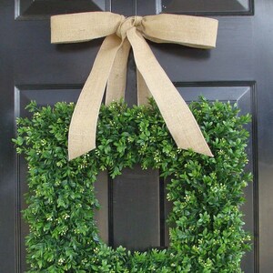 May include: A square-shaped boxwood wreath with a burlap bow hanging on a black door. The wreath is made of green boxwood leaves and has a square opening in the center.