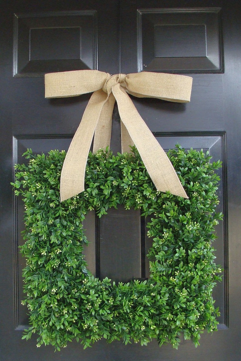 May include: A square-shaped boxwood wreath with a burlap bow hanging on a black door. The wreath is made of green boxwood leaves and has a square opening in the center.