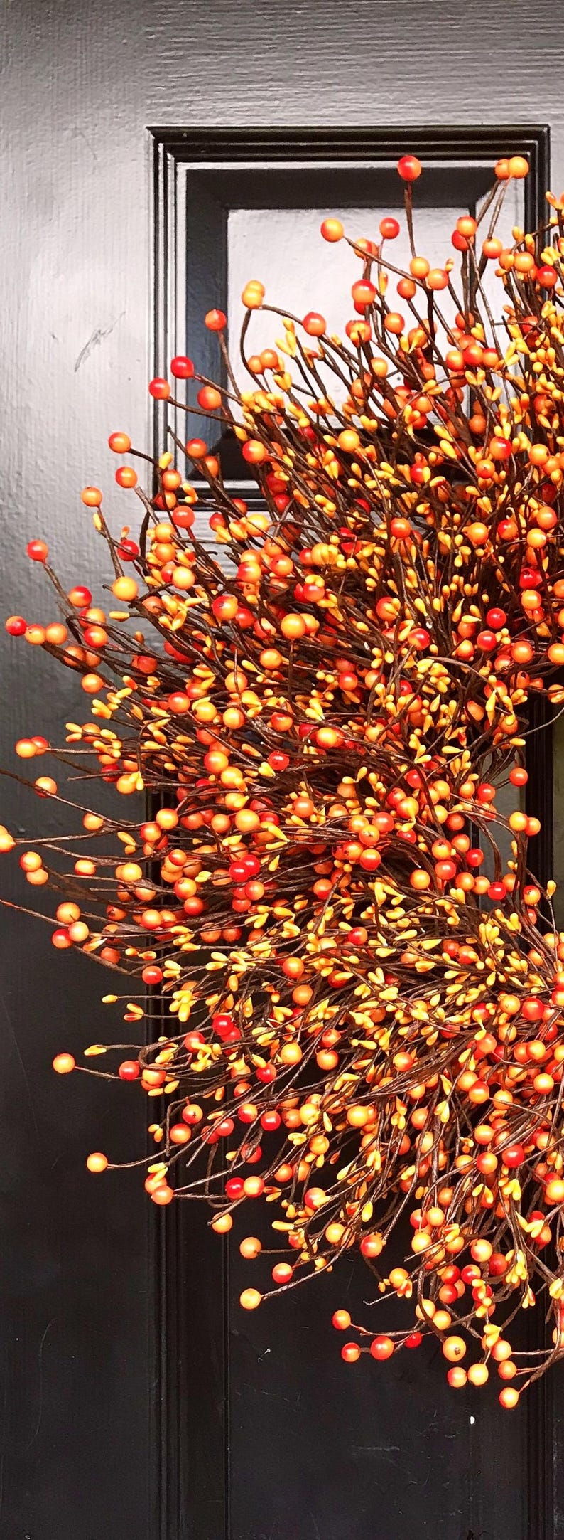 Puede incluir: Una corona hecha de ramas marrones con peque&ntilde;as bayas naranjas y rojas. La corona est&aacute; colgada en una puerta negra.