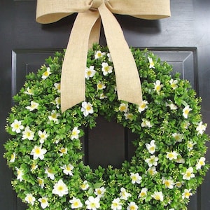 May include: A green wreath with white flowers and a burlap bow. The wreath is hanging on a black door.