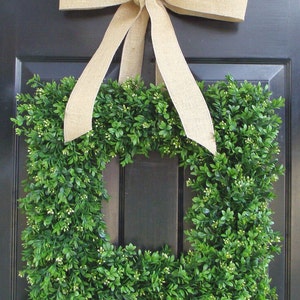 May include: A square boxwood wreath with a burlap bow hanging on a black door. The wreath is made of green boxwood leaves and has a square shape.