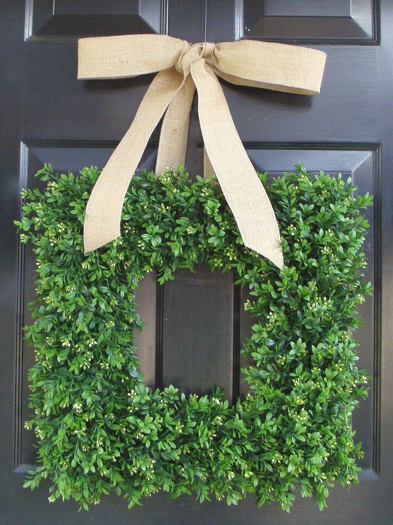 May include: A square boxwood wreath with a burlap bow hanging on a black door. The wreath is made of green boxwood leaves and has a square shape.