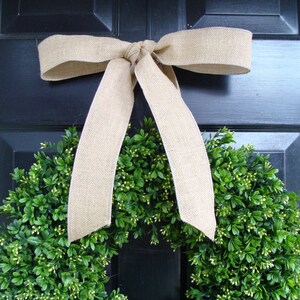 May include: A green boxwood wreath with a burlap bow hanging on a black door.