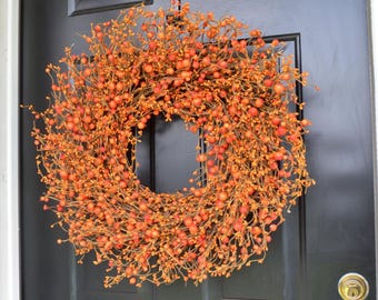 May include: A circular wreath composed of numerous small, round orange and red berries attached to thin, branching stems. The wreath is mounted on a solid black door featuring rectangular panels and a metallic gold-colored circular door handle.