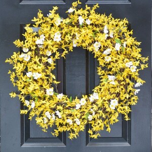 May include: A yellow and white floral wreath with small white flowers and yellow leaves hangs on a black door.
