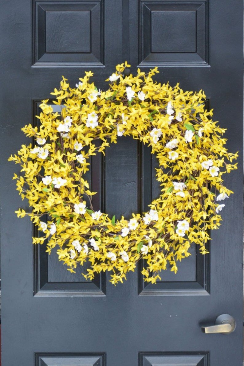 May include: A yellow and white floral wreath with small white flowers and yellow leaves hangs on a black door.