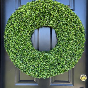 May include: A vibrant green wreath made of artificial boxwood foliage hangs on a black front door. The wreath is circular, with a dense, textured appearance. The door has a glossy finish and a simple, elegant design.