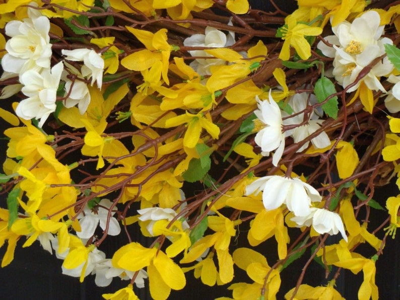 May include: A wreath made of artificial yellow and white flowers. The flowers are arranged in a circular shape and are attached to brown branches.