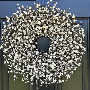 May include: A circular wreath constructed from thin, dark brown intertwined branches densely covered in numerous small, matte white berries. The arrangement features a full, rounded shape with a hollow centre, creating a textured, spherical appearance.